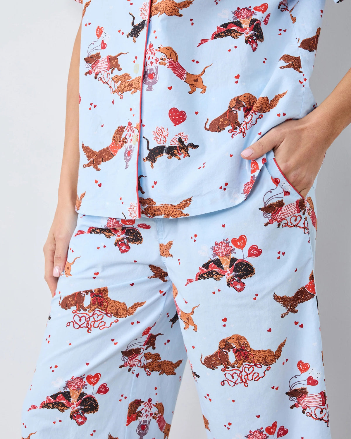 A woman with long, wavy brown hair stands barefoot against a white wall, wearing the PRINTFRESH Dachshund Date Short Sleeve Top & Cropped Pants Pajama Set in Blue. The organic cotton set from PRINTFRESH features short sleeves and cropped pants adorned with a charming dachshund print. She smiles slightly.