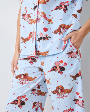 A woman with long, wavy brown hair stands barefoot against a white wall, wearing the PRINTFRESH Dachshund Date Short Sleeve Top & Cropped Pants Pajama Set in Blue. The organic cotton set from PRINTFRESH features short sleeves and cropped pants adorned with a charming dachshund print. She smiles slightly.