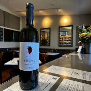 A bottle of ENATE CABERNET SAUVIGNON 2016 by OCEAN SOUTH IMPORTS, a distinguished Somontano wine aged in French oak, stands on a reflective bar counter in a modern restaurant, with menus and a vase of flowers in the background.