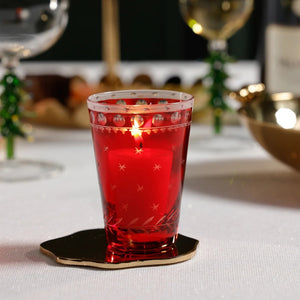 A ZODAX HAND CUT TEA LIGHT CUP, a red borosilicate glass votive holder with etched star and circle patterns, containing a lit candle and sitting on a gold-toned coaster.