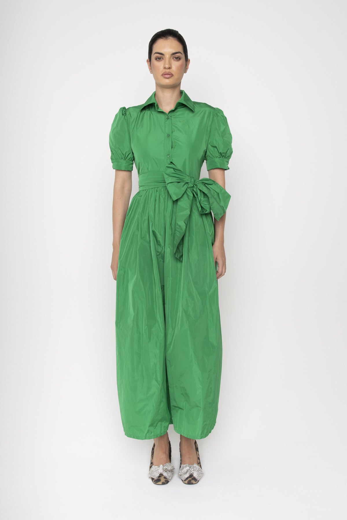 A person stands against a plain background wearing the vibrant green LAVI COUTURE Salcerella Dress, featuring puffed sleeves and a large waist bow. Paired with patterned high heels, this Italian-made piece exudes elegance.