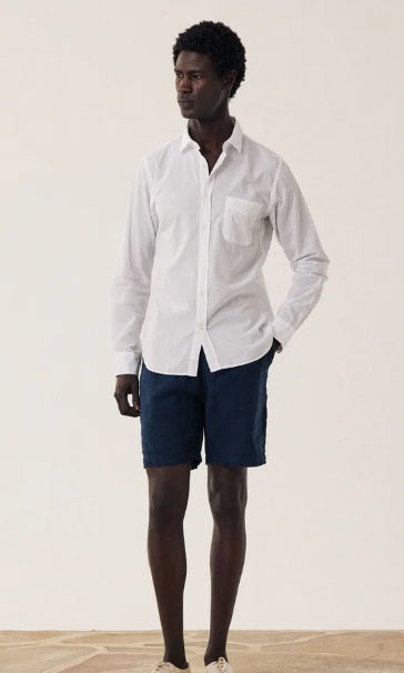 A person wearing a white long-sleeve shirt, HARTFORD - LINEN JOGGY SHORT IN NAVY by HARTFORD, and beige sneakers stands on a wooden floor against a plain light background.