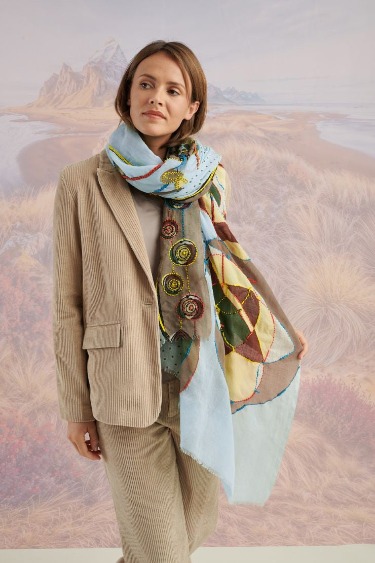 A woman with long blonde hair smiles at the camera in a scenic setting, wearing a white shirt, plaid pants, and the STORIATIPIC - ASTER WOOL SCARF by STORIATIPIC. Mountains, grass, and water are visible in the background.