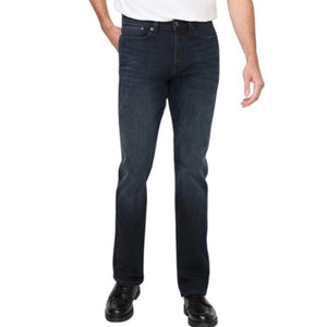 A man stands against a plain background wearing a black jacket, white t-shirt, DU/ER - PERFORMANCE DENIM+ SLIM STRIAGHT PANTS IN CARBON STONE, and black shoes, with one hand in his pocket and a neutral expression.