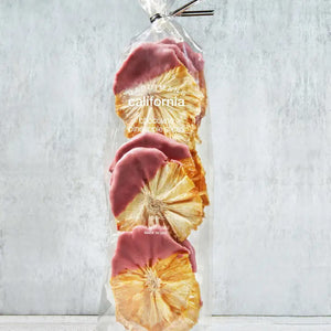 A clear plastic bag from DARDIMANS CALIFORNIA CRISPS contains several dried pineapple slices that are half-dipped in ruby chocolate. The label on the bag reads "DARDIMANS - RUBY CHOCOLATE PINEAPPLE SLICES." The bag, brimming with tropical sweetness, is secured with a twist tie and set against a textured, light grey background.