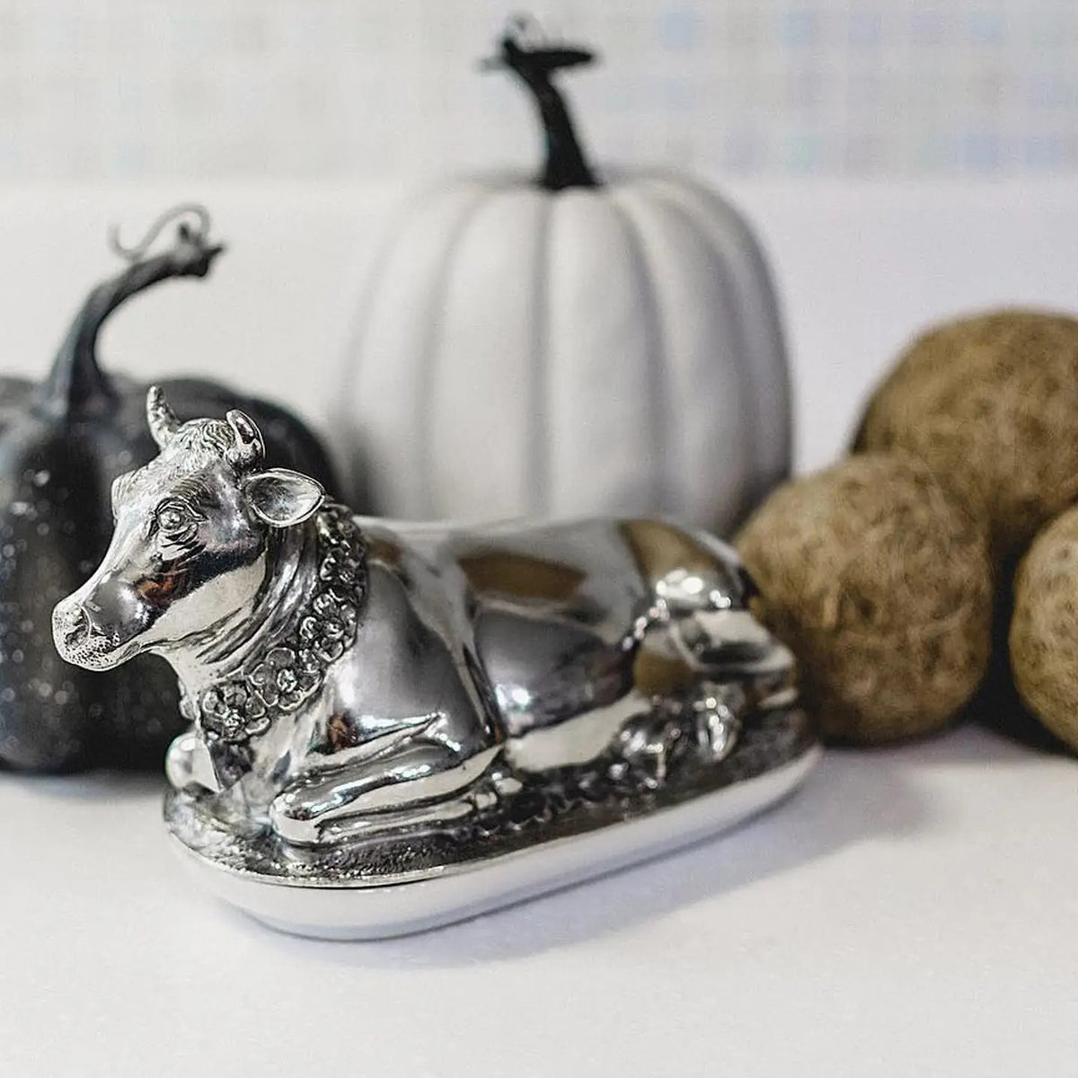 The VAGABOND HOUSE Mabel Cow Butter Dish, by VAGABOND HOUSE, is a silver-toned luxury pewter piece with intricate details. The cow’s textured features and elegant oval base highlight its exquisite craftsmanship.