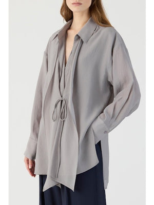 A woman wears the ELEH - BLOUSE WITH DRAPE FRONT in light gray, featuring relaxed tailoring, long sleeves, a collar, front tie detail, and an asymmetrical hem. It pairs effortlessly with dark pants for a chic look.