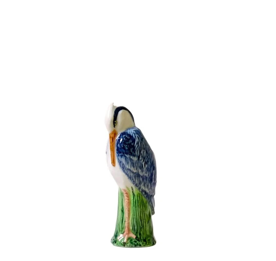 The QUAIL CERAMICS - HERON BUD VASE by QUAIL DESIGNS features a hand painted stoneware heron with blue and white feathers, orange beak, and green legs perched on a green base, shown against a plain white background.