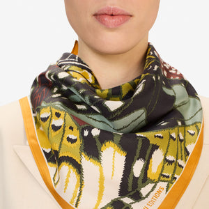 A woman with dark hair wears a beige blazer and the INOUI EDITIONS - PAPILLON (BUTTERFLIES) Silk Scarf in Green by INOUI, a versatile accessory made in India, featuring vibrant yellow, black, and green designs, against a plain white background.