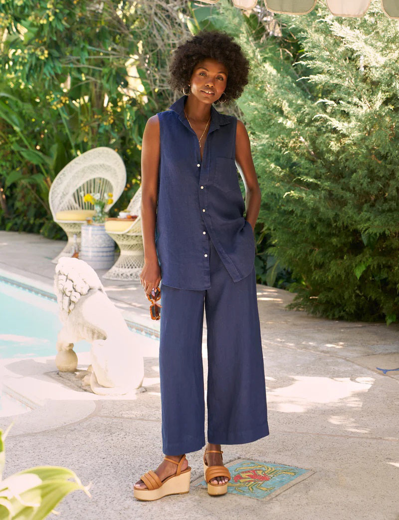 A woman with curly hair stands by a pool, wearing the FRANK &amp; EILEEN Finley Sleeveless Button Up Shirt in washed linen navy and matching wide-leg pants with tan platform sandals. Lush greenery and outdoor furniture are behind her.