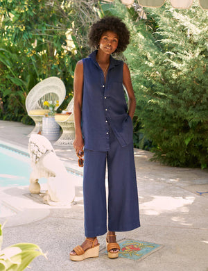 A woman with curly hair stands by a pool, wearing the FRANK & EILEEN Finley Sleeveless Button Up Shirt in washed linen navy and matching wide-leg pants with tan platform sandals. Lush greenery and outdoor furniture are behind her.