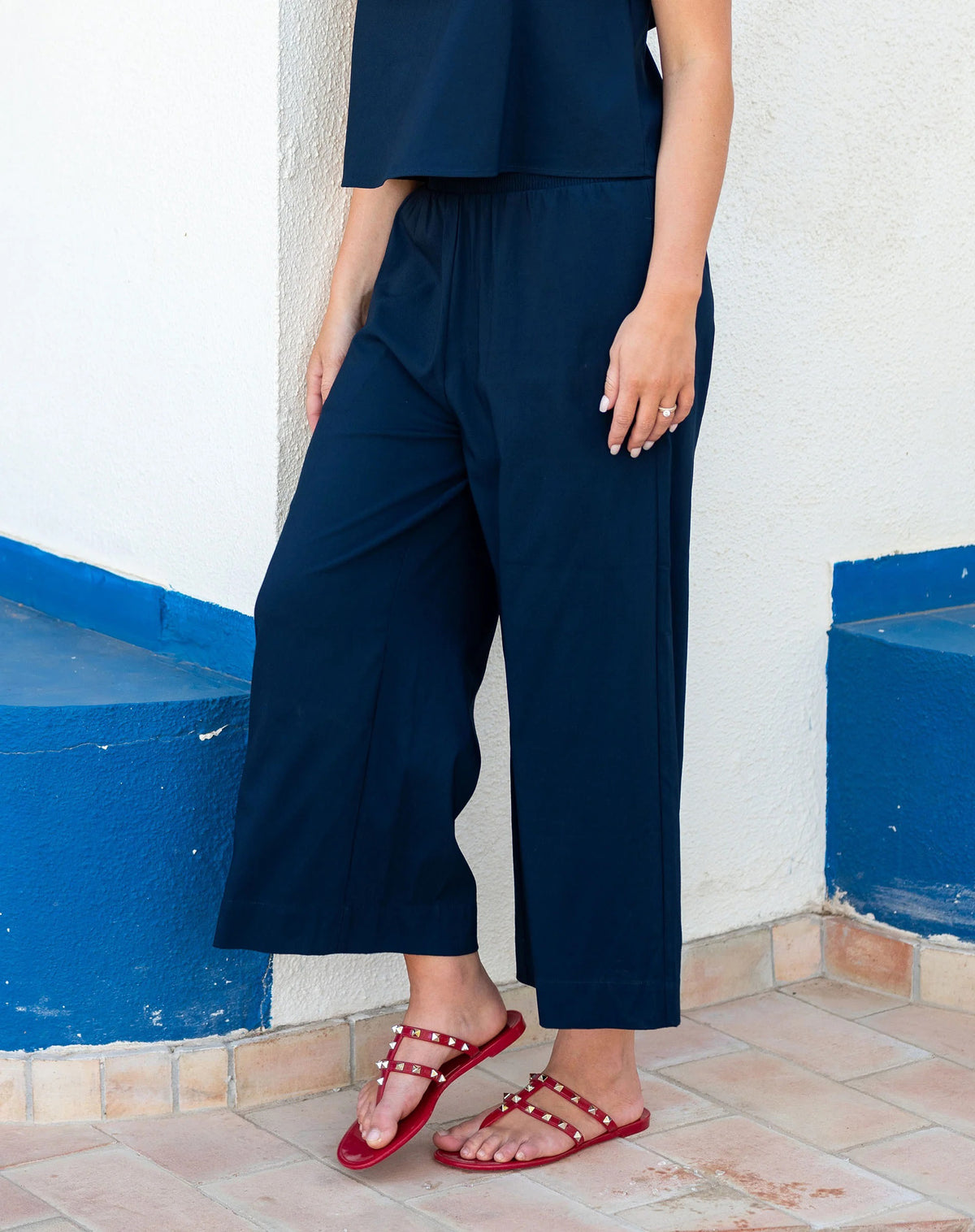 A person stands on tiled flooring wearing the MERSEA - VERANDA CROPPED PANT in navy blue with a ruffle waistband, a matching top, and red studded sandals. Their left hand shows a ring. The white wall behind them has blue accents.