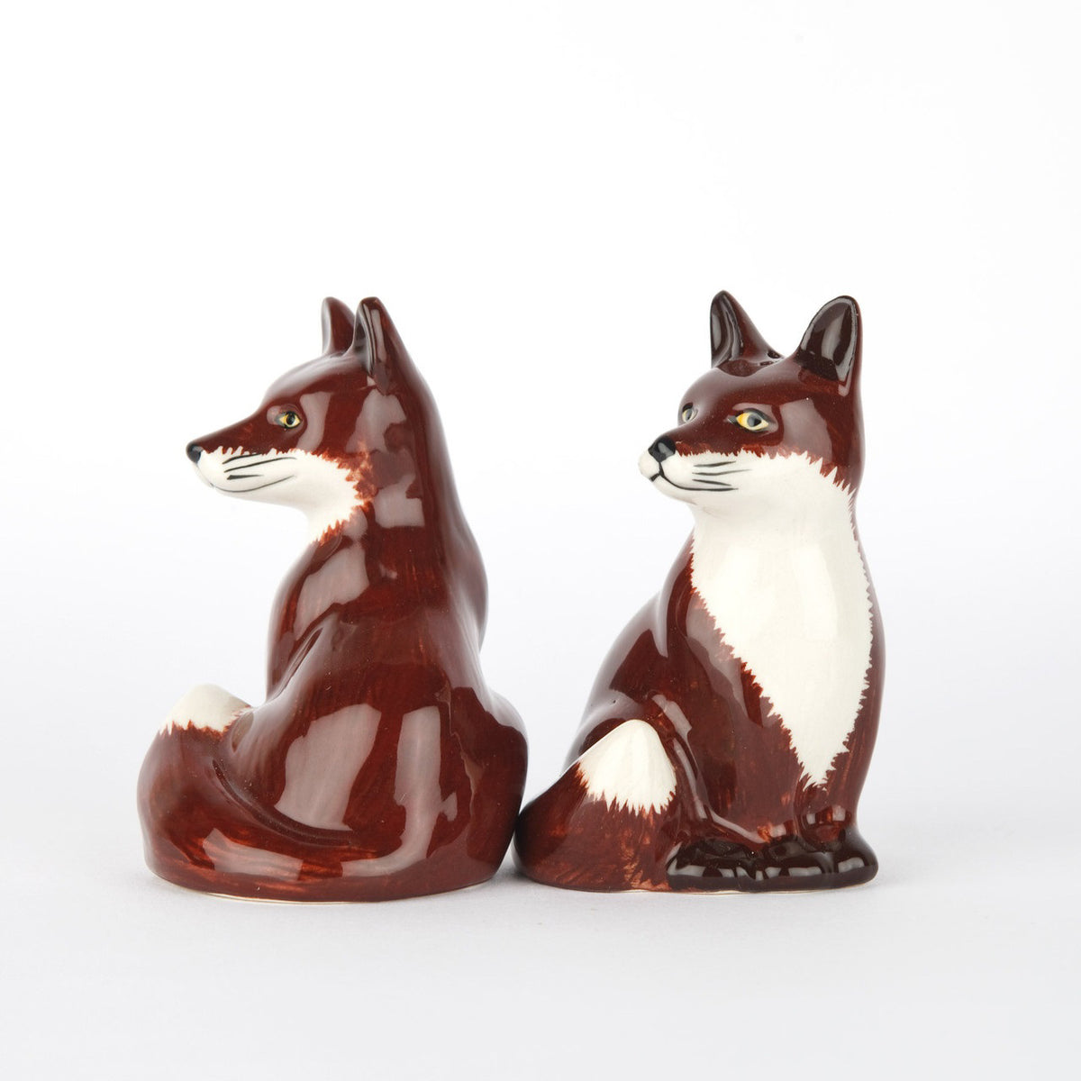The QUAIL DESIGNS QUAIL CERAMICS - FOX SALT AND PEPPER SET features two handpainted stoneware shakers with reddish-brown and white fur, upright ears, bushy tails, and alert expressions, shown side by side on a white background.