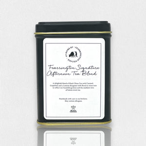 A black tin labeled "Fearrington Signature Afternoon Tea Tin" from the Fearrington House Signature Collection contains a luxurious blend of Loose Leaf Black China Tea with caramel, grapefruit, and lavender. The tin has a simple, elegant design accented by a white label featuring the product description.