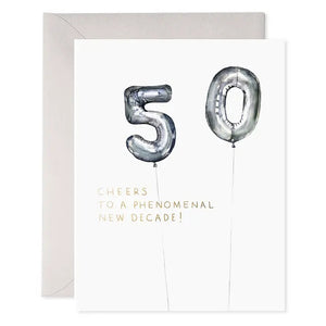 The E. FRANCES PAPER Helium 50 Birthday Card features silver balloon numbers "50" and the phrase "Cheers to a phenomenal new decade!" in gold, making it the perfect festive balloon birthday card. A light grey envelope is positioned behind the card, set against a clean white background.