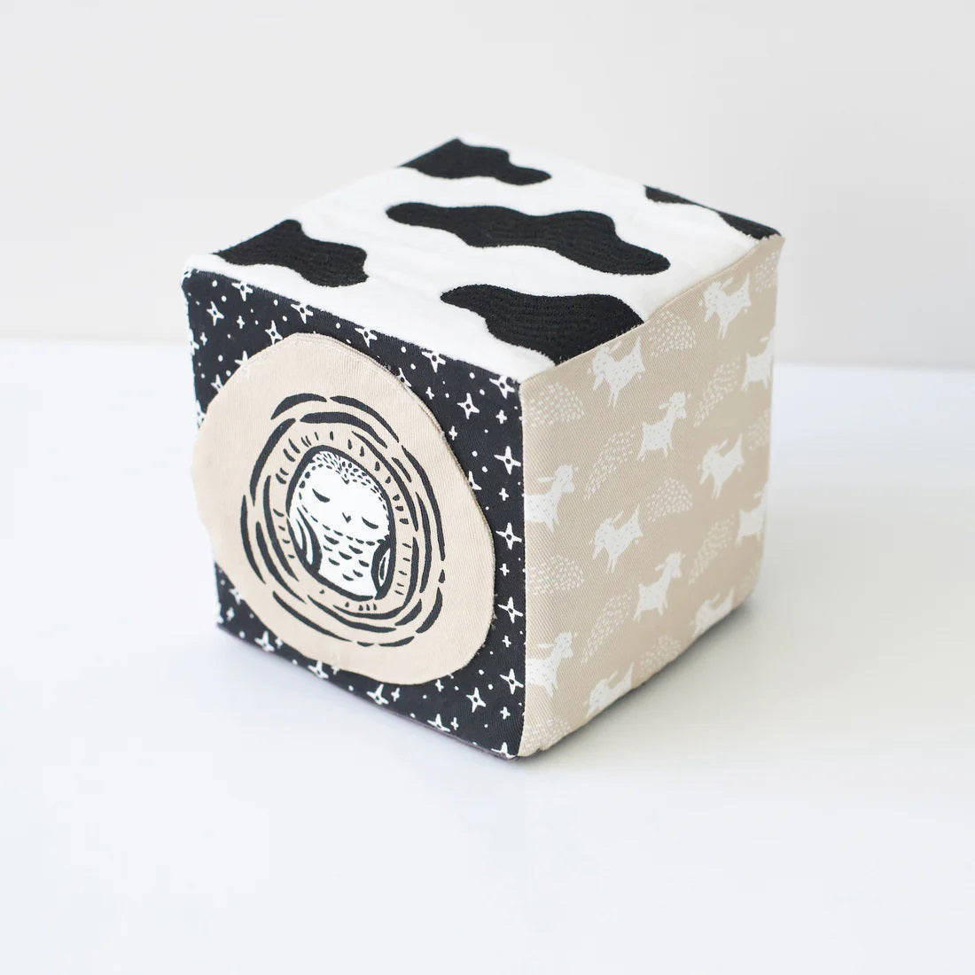 The WEE GALLERY - BABY ANIMALS SOFT BLOCK by WEE GALLERY is a fabric cube adorned with black and white designs that include adorable baby animals, such as a sleeping fox and its cub on one side. This sensory development toy features various textures, including polka dots and stripes, and includes two fabric loops on one edge to aid in tactile exploration.