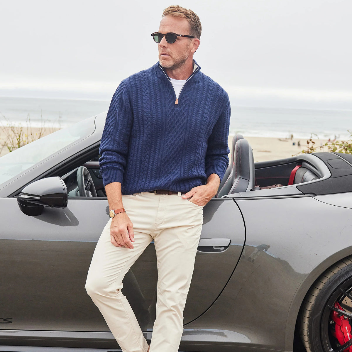 A man in COLLARS AND CO’s Napoli Cable Knit Quarter Zip Wool and Cashmere Sweater in Navy stands by a convertible near the beach, with the ocean and a cloudy sky behind him.