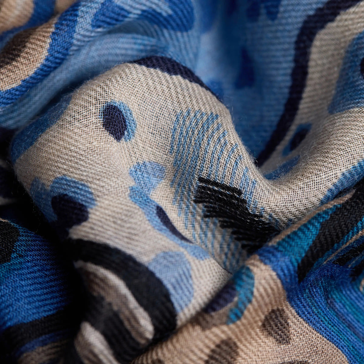 A person with straight, reddish-brown hair models the INOUI EDITIONS - WESTERN SQUARE SCARF by INOUI, a blue and white patterned wool accessory made in India, draped over a light gray ribbed sweater and black pants, standing against a plain white background.