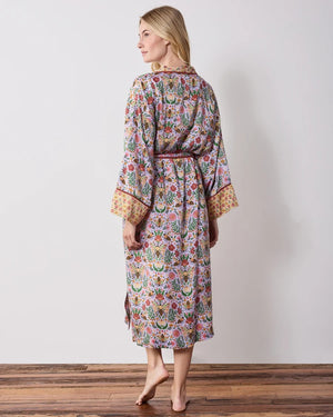 A woman wears a PRINTFRESH - BEE TAPESTRY ECO SATIN ROBE IN LAVENDER, featuring a lavender floral-print with a repeating pattern of bees, flowers, and vines with a contrasting yellow and red patterned trim.