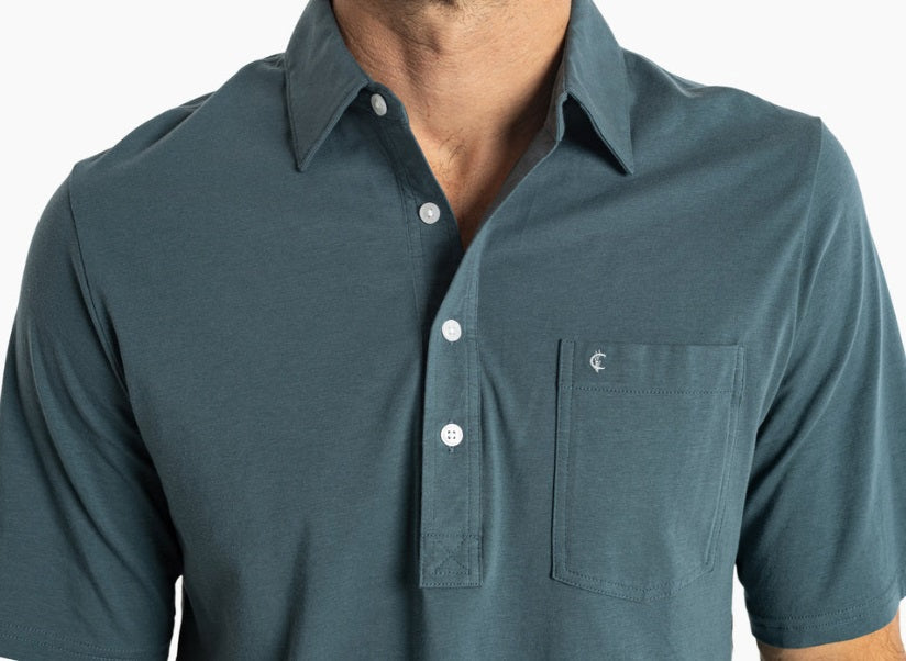 A smiling man with short dark hair wears the CRIQUET Top-Shelf Players Shirt in Dark 'n Stormy with a chest pocket and dark jeans, standing against a plain white background.