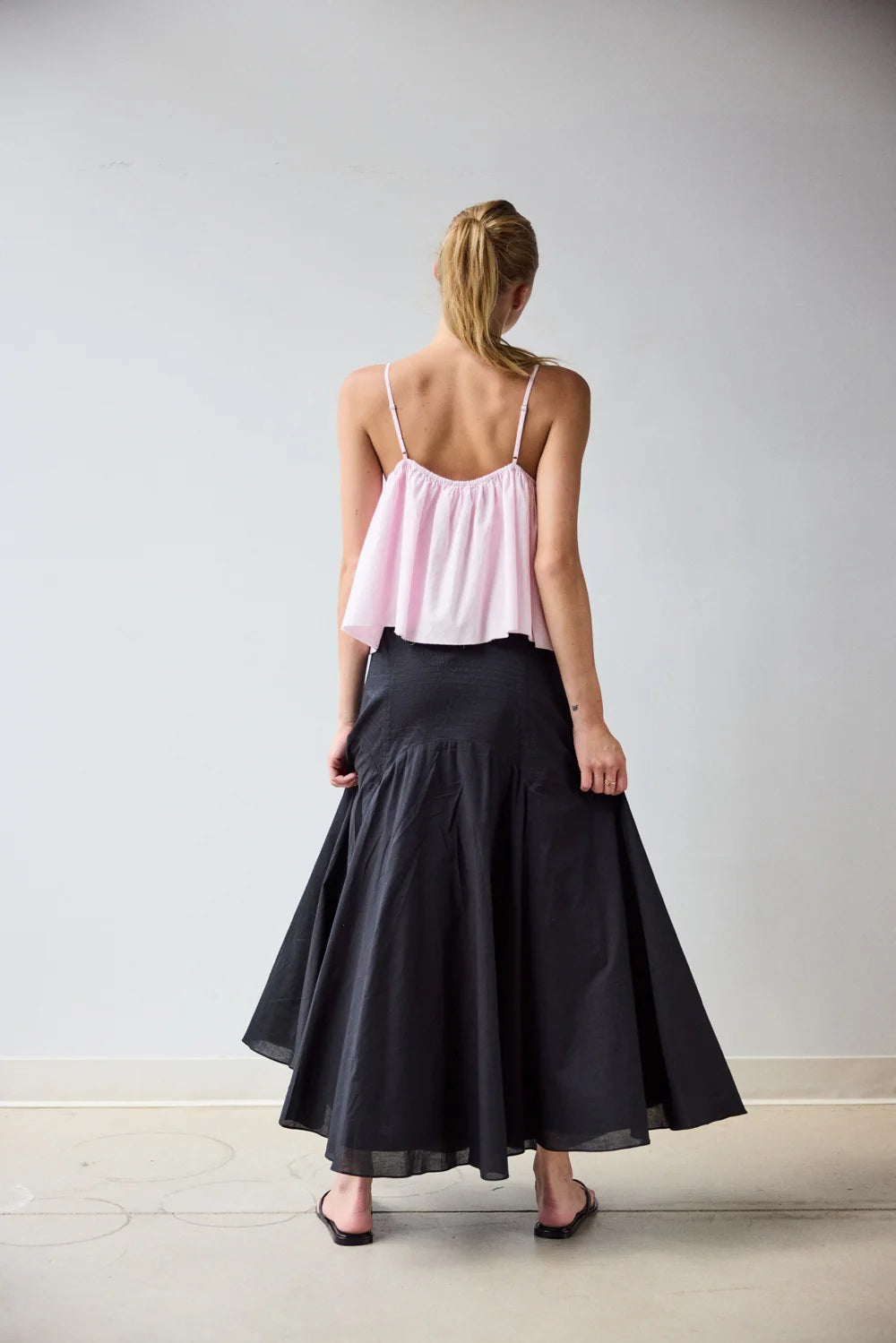 A woman with blonde hair tied back stands barefoot on a light floor, wearing a light pink spaghetti strap top and the HOPE FOR FLOWERS - CIRCLE SKIRT IN BLACK, made from GOTS Certified organic cotton, facing away against a plain wall.
