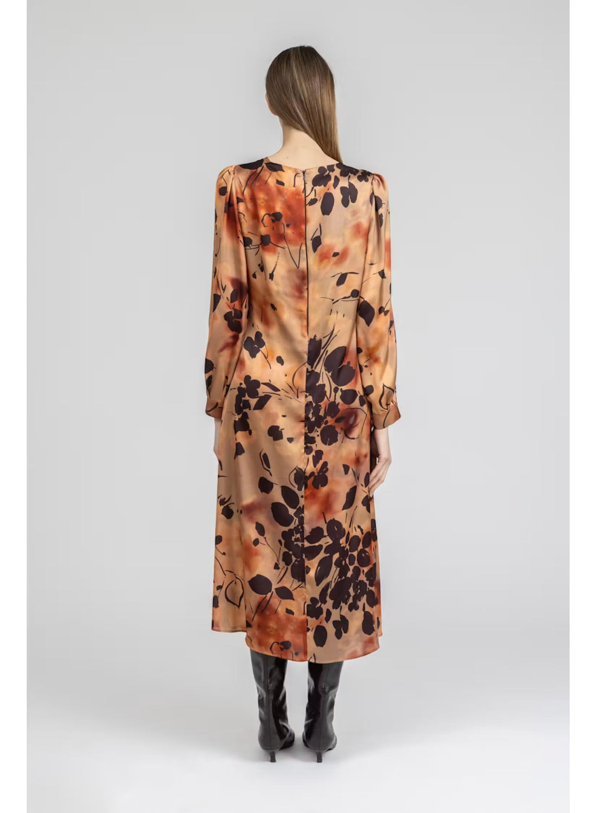 A woman stands facing forward against a plain light gray background, wearing the MILVA MILVA MI - BURNT ORANGE PRINTED DRESS with long sleeves and an autumn-inspired floral pattern, styled with black knee-high boots.