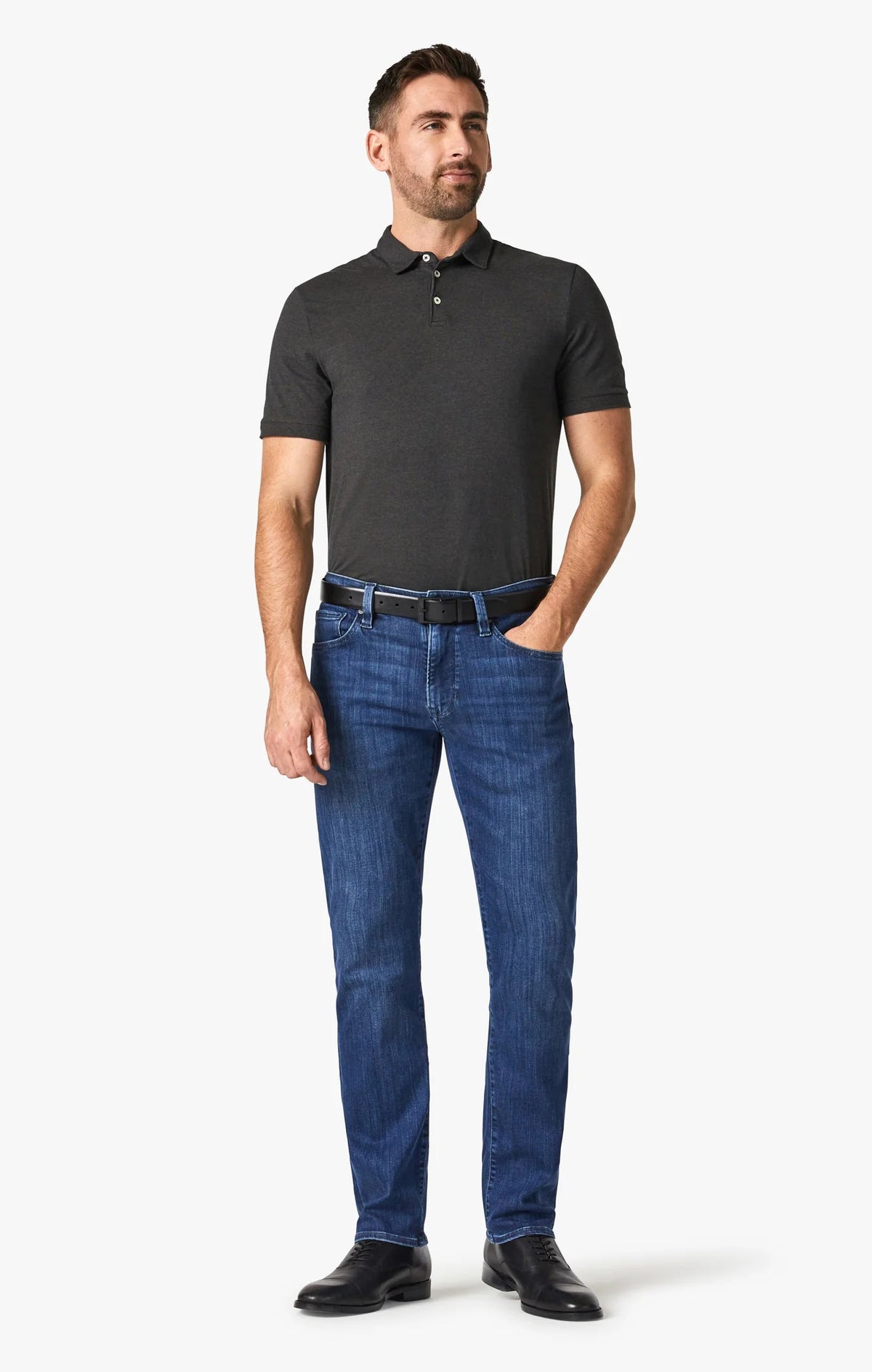 A man stands facing forward, wearing a dark short-sleeve polo, 34 HERITAGE - COURAGE STRAIGHT LEG JEAN IN MID URBAN WASH, a black belt, and black shoes. One hand is in his pocket; he has short hair, a trimmed beard, and a neutral expression.