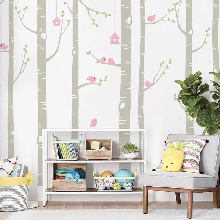 A whimsical baby nursery features a wall adorned with SIMPLE SHAPES - BIRCH TREE BIRDS REMOVABLE WALL DECALS from SIMPLE SHAPES, showcasing birch trees and cute pink birds. Below the mural, a white storage shelf holds books and toys. A cushioned chair with a striped pillow and a plant add warmth to the cozy space.