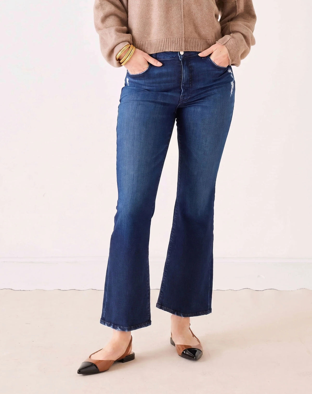 A person stands with hands in pockets against a plain light background, wearing MERSEA’s Murphy Mini Boot Jeans in Patriot—a pair of mid-rise, blue flare jeans crafted from four-way stretch denim—paired with a tan long-sleeve top, black pointed-toe flats, and gold bangles.