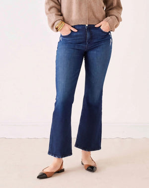 A person stands with hands in pockets against a plain light background, wearing MERSEA’s Murphy Mini Boot Jeans in Patriot—a pair of mid-rise, blue flare jeans crafted from four-way stretch denim—paired with a tan long-sleeve top, black pointed-toe flats, and gold bangles.