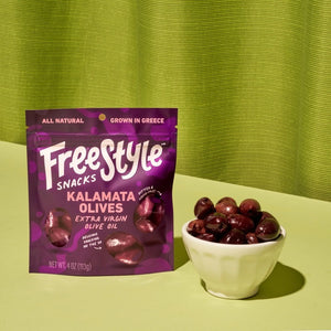 The purple FREESTYLE SNACKS package of seasoned Kalamata olives in extra virgin olive oil says "Olives, Reimagined." It offers non-GMO, gluten-free nutrition details and suggests pairings like hummus and goat cheese. Enhanced with sea salt, it includes a resealable opening.
