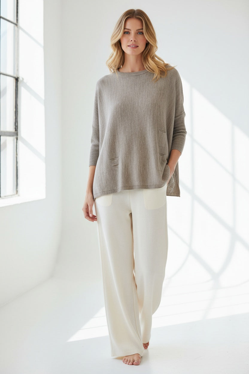 A woman with curly hair wears the MERSEA - CATALINA CREWNECK SWEATER IN DRIFTWOOD, a relaxed fit sweater by MERSEA, layered over a light skirt. She smiles gently while looking down, one hand touching her hair and the other resting on her wrist. The background is plain white.
