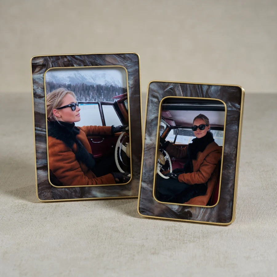 A person wearing sunglasses and a brown coat sits in a vintage car, holding the steering wheel. The photo is displayed in the ZODAX Marbled Hazel Resin Frame with Antique Brass Trim (4x6), featuring gold accents.