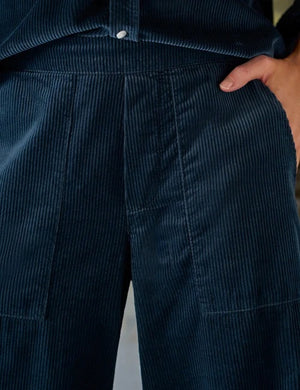 A person stands indoors by a window, dressed in the FRANK & EILEEN MADELEINE Patch Pocket Pull On Pant in Italian Corduroy Classic Navy and a coordinating dark navy button-up shirt, paired with brown shoes. With one hand tucked into their pocket, they smile softly as sunlight streams into the minimalist room.