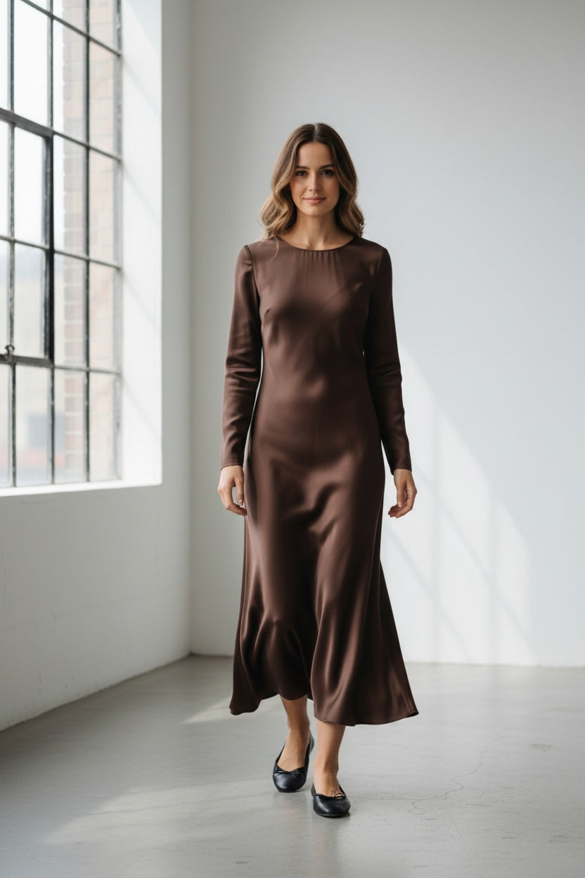 A woman with long blonde hair models the MILVA MI - SATIN DRESS IN MORRO by MILVA, an elegant chocolate brown satin dress featuring long sleeves and an ankle-length silhouette, paired with black flats against a plain light-colored background.