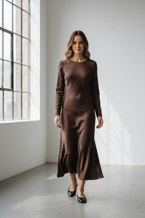 A woman with long blonde hair models the MILVA MI - SATIN DRESS IN MORRO by MILVA, an elegant chocolate brown satin dress featuring long sleeves and an ankle-length silhouette, paired with black flats against a plain light-colored background.