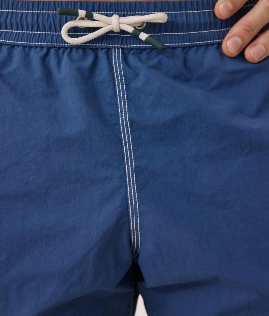 A shirtless person models the HARTFORD - NYLON SWIM TRUNKS WITH TRAVEL POUCH IN DENIM by HARTFORD against a plain light background, with tattoos visible on their arms and legs and one hand casually in a pocket.