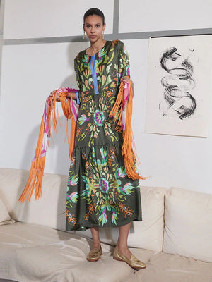 A woman wears the FRANCO FERRARI - SILK TIERED MIDI DRESS, a long sleeve olive green silk piece with colorful floral and abstract prints. She smiles softly, has wavy hair, beige sandals, and stands against a plain light gray background.