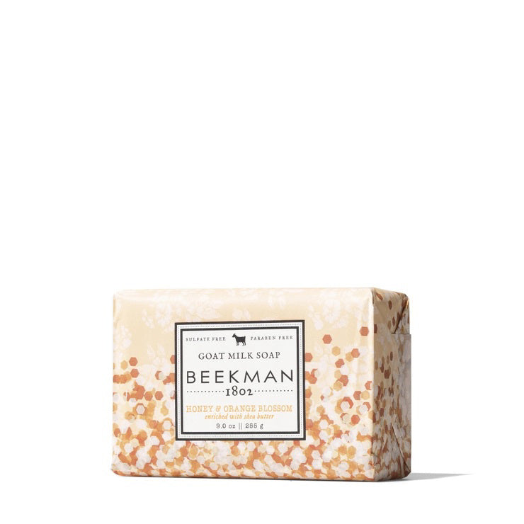 A bar of BEEKMAN - 9OZ. HONEY &amp; ORANGE BLOSSOM BAR SOAP from the BEEKMAN - STA ELEMENTS line is wrapped in decorative paper adorned with white and orange floral patterns. The label highlights that it is free from sulfates and parabens, featuring an illustration of a goat above the brand name, assuring you it&#39;s a moisturizing soap you can trust.