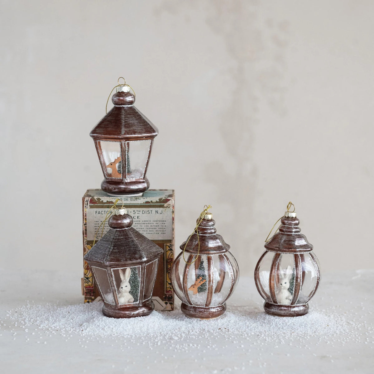 Four CREATIVE COOP GLASS LANTERN WITH WOODLAND ANIMALS ornaments—two with bunnies and two with birds—rest on faux snow before a decorative box, all set against a softly textured, neutral backdrop.