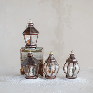 Four CREATIVE COOP GLASS LANTERN WITH WOODLAND ANIMALS ornaments—two with bunnies and two with birds—rest on faux snow before a decorative box, all set against a softly textured, neutral backdrop.