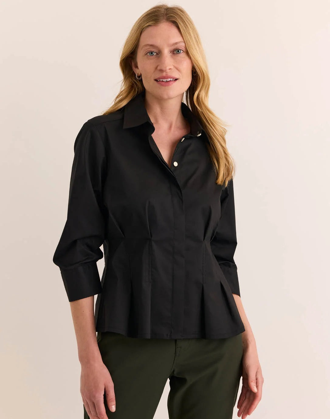 A woman with blonde hair is smiling at the camera while standing against a plain light background, wearing the HINSON WU - MARTINA 3/4 SLEEVE STRETCH POLISHED COTTON SHIRT IN BLACK paired with olive green pants.