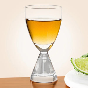 The ZODAX TIMELESS SIP SHOT GLASS, featuring a unique double-ended shape, sits half-filled with amber liquid on a light surface, while two dark round dishes are softly blurred in the background.