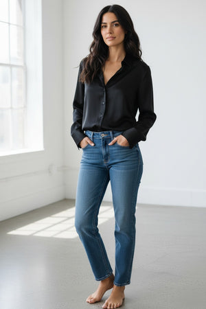 A woman with long dark hair is wearing the HINSON WU Lindsey Long Sleeve Liquid Satin Shirt in Black, which features a relaxed fit, paired with blue jeans. She stands against a plain light-colored background, looking at the camera with a slight smile.