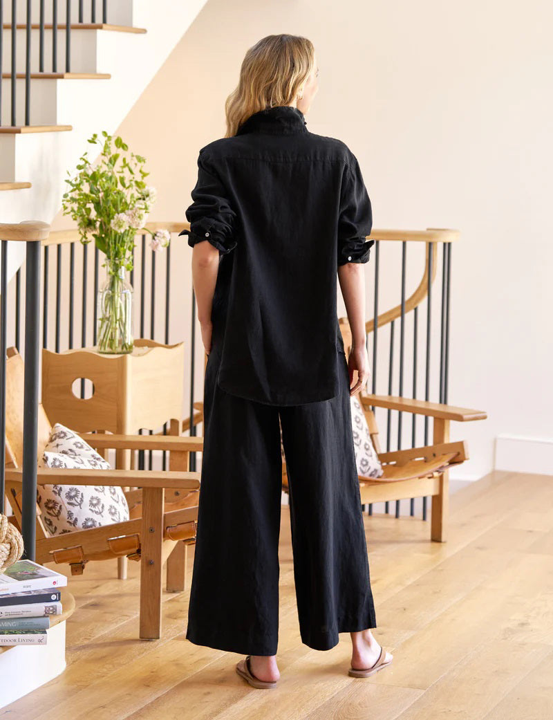 A woman with wavy blonde hair stands indoors in a loose black shirt and FRANK &amp; EILEEN’s Maisie Wide Leg Pull On Pant in Washed Linen Black, barefoot on a wooden floor near a chair, vase of green flowers, and staircase.