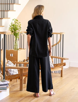 A woman with wavy blonde hair stands indoors in a loose black shirt and FRANK & EILEEN’s Maisie Wide Leg Pull On Pant in Washed Linen Black, barefoot on a wooden floor near a chair, vase of green flowers, and staircase.