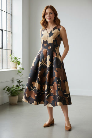 A woman stands with her hands in the pockets of the PARVATI DRESS by ALYKI LABORATORIO, a sleeveless, V-neck, calf-length dress featuring a brown and black abstract floral pattern, styled with black knee-high boots.
