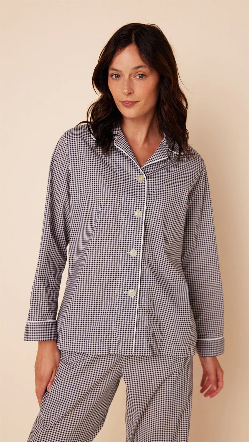 A person with long dark hair is standing on a light beige background, wearing a matching set of loose-fitting, long-sleeved CAT&#39;S PAJAMAS - CLASSIC GINGHAM LUXE PIMA PAJAMA by CAT&#39;S PAJAMAS. Made from Luxe Pima Cotton, the buttoned-up pajamas feature a collar. The person is barefoot and looking slightly towards the camera.