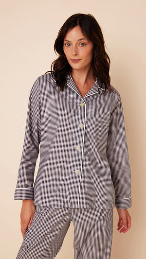 A person with long dark hair is standing on a light beige background, wearing a matching set of loose-fitting, long-sleeved CAT'S PAJAMAS - CLASSIC GINGHAM LUXE PIMA PAJAMA by CAT'S PAJAMAS. Made from Luxe Pima Cotton, the buttoned-up pajamas feature a collar. The person is barefoot and looking slightly towards the camera.
