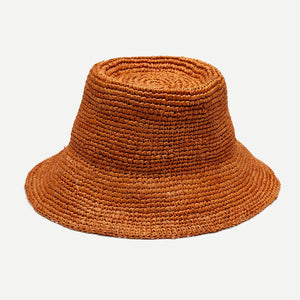 A woman with long, wavy blonde hair wears the WYETH TALI Raffia Bucket Hat in Rust and a white sleeveless top, standing in profile against a plain light background.